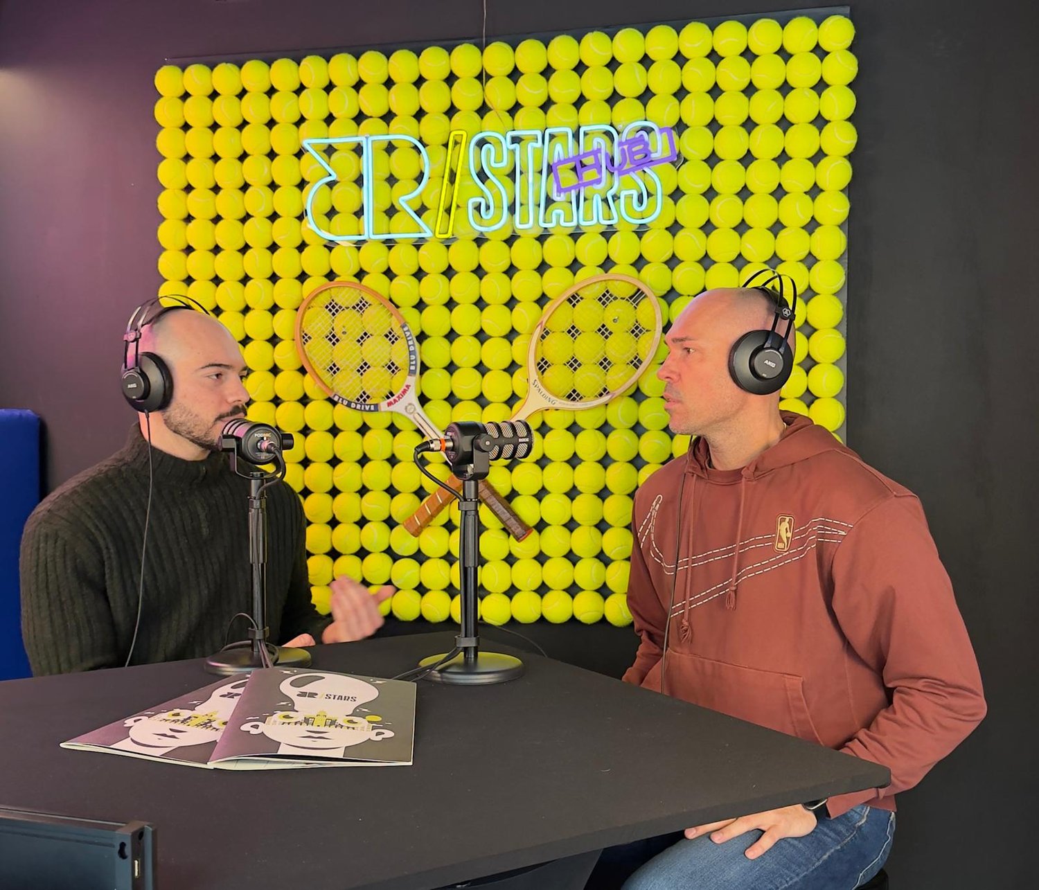 Due uomini con cuffie registrano un podcast davanti a una parete di palline da tennis gialle con neon R/STARS LIVE